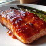 Close-up of a perfectly cooked fillet of Spicy Salmon coated in a thick, glossy red glaze, served next to roasted asparagus.