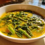 Close-up of a white bowl filled with vibrant yellow Spring Detox Soup with Ginger Turmeric, featuring bright green asparagus and leafy greens.
