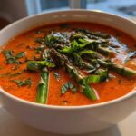 Close-up of a bowl of vibrant orange Spring Soup with Roasted Tomatoes, topped with bright green asparagus spears and fresh basil.