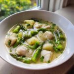 A white bowl filled with vibrant Spring Soup with White Fish, featuring chunks of white fish, green asparagus, and potatoes in a clear broth.