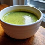 Close-up of a bowl filled with bright green Spring Soup with Zucchini and Basil, resting on a wooden surface near a window.