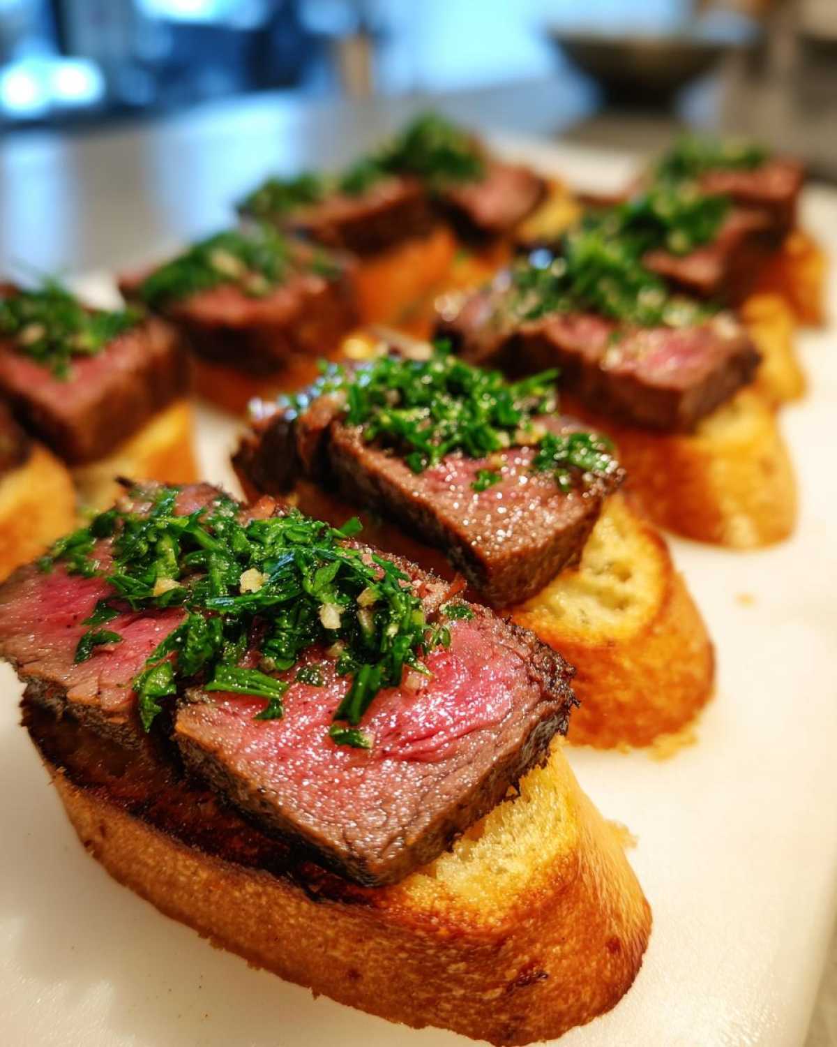 Close-up of several steak crostini appetizers topped with medium-rare steak slices and green herb topping.