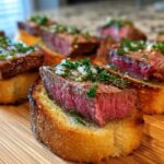 Close-up of several steak crostini appetizers featuring medium-rare steak slices topped with herbs and sea salt on toasted bread.