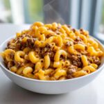 Close-up of a white bowl filled with hot, steaming cheeseburger pasta featuring elbow macaroni and seasoned ground beef in a cheesy sauce.