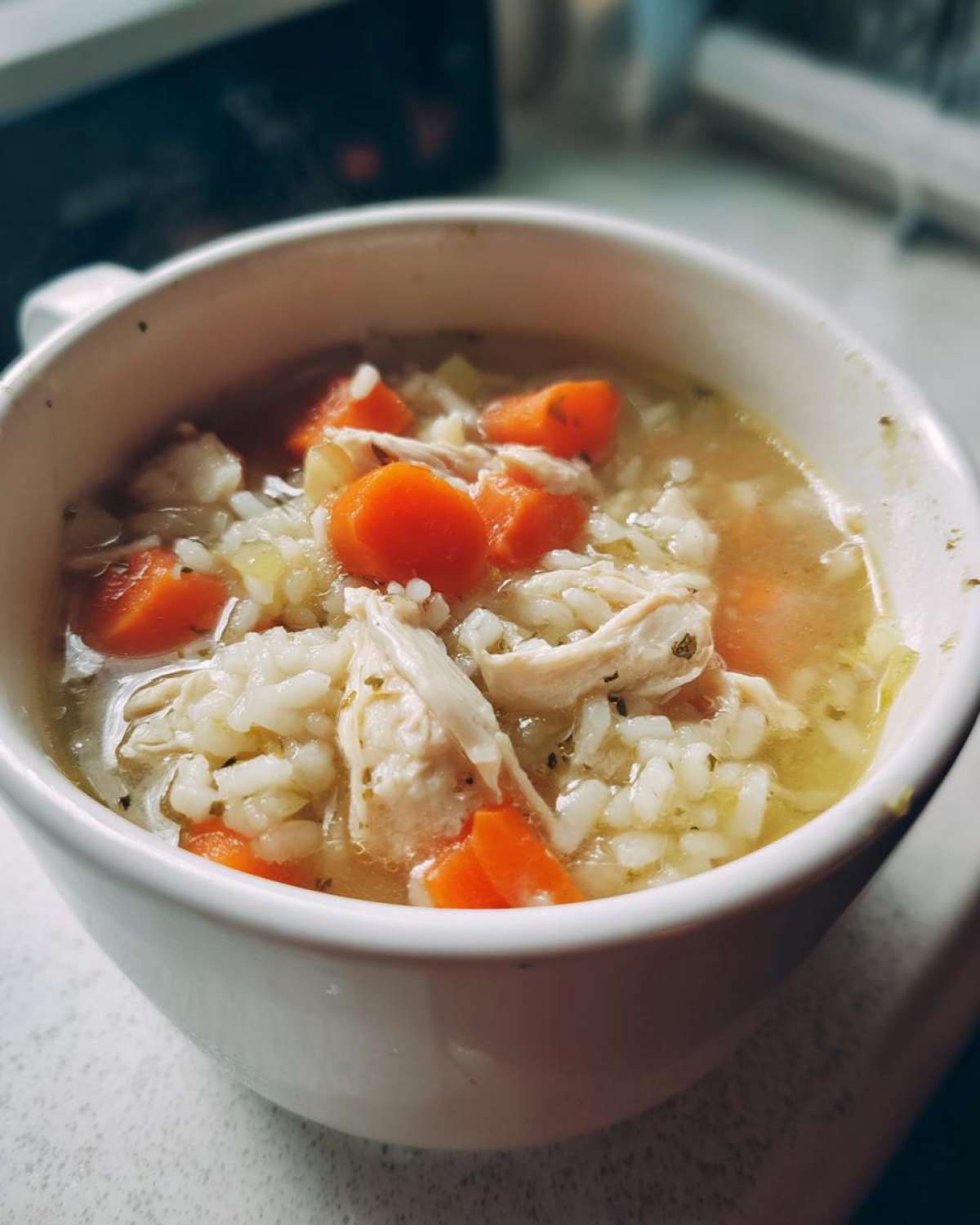 Close-up of a steaming white bowl filled with homemade chicken and rice soup, featuring shredded chicken and bright orange carrots.