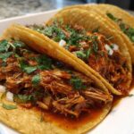 Close-up of three saucy chicken tacos filled with shredded chicken, cilantro, and onion, resting in a rich sauce.
