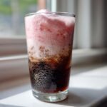 A tall glass of cold brew coffee topped with thick, pink strawberry cold foam, condensation visible on the glass.