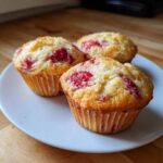 Three golden-brown Strawberry Yogurt Muffins studded with bright red fruit pieces, resting on a white plate.