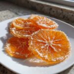 Close-up of several bright orange candied orange slices heavily coated in sparkling sugar crystals on a white plate.