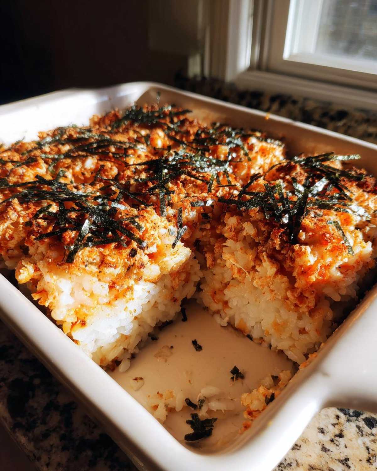 A square white baking dish filled with golden brown sushi bake casserole, showing a piece removed, topped with crispy flakes and shredded nori.