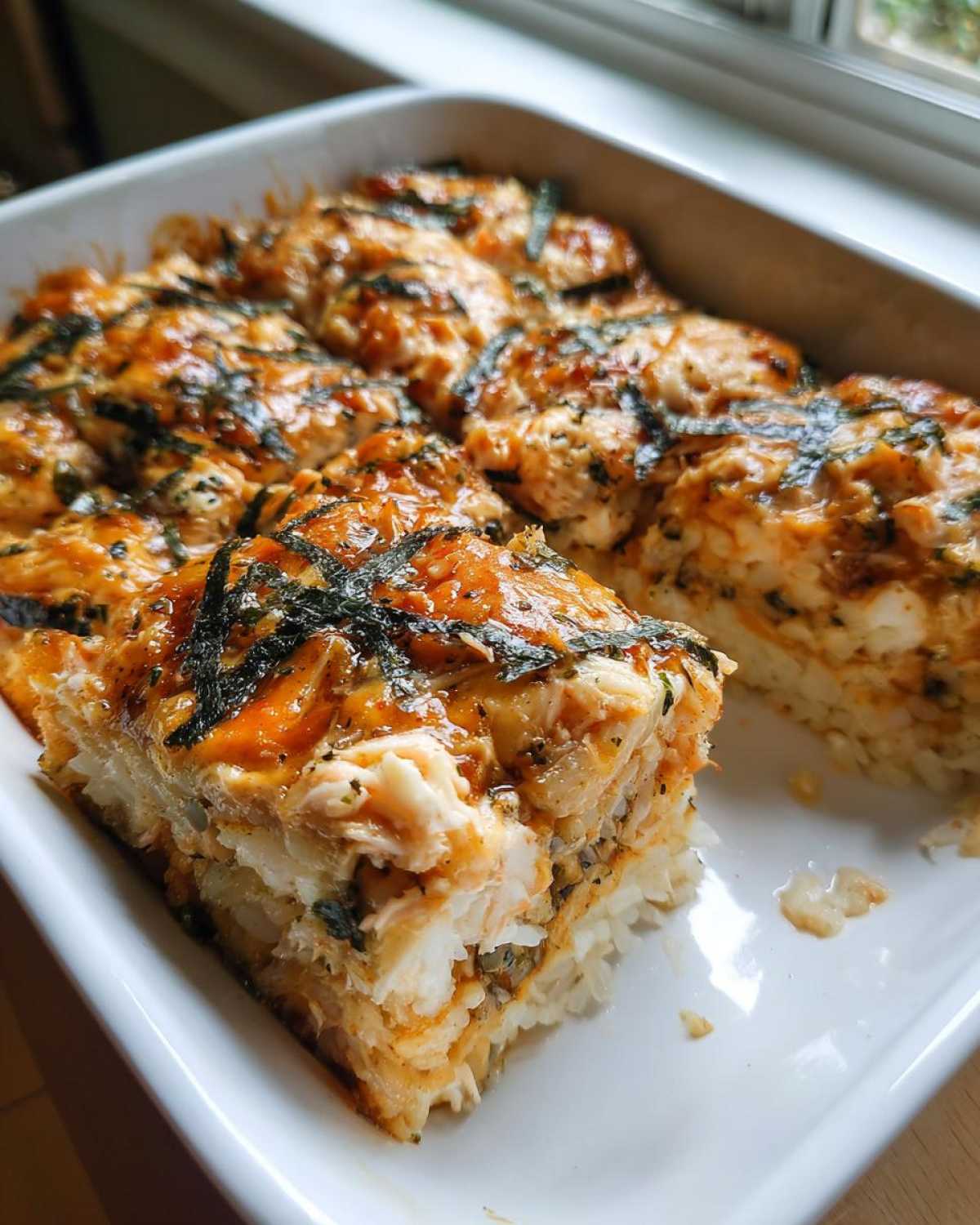 A close-up of a rich, baked sushi bake casserole in a white dish, showing a cut portion revealing layers of rice and creamy topping.