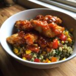 A close-up of a bowl featuring breaded chicken pieces covered in a glossy, sweet and spicy sauce over a bed of quinoa mixed with corn and peppers.