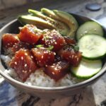 A close-up view of a fresh Tuna Poke Bowl featuring marinated tuna cubes over rice, topped with sesame seeds, sliced avocado, and cucumber.