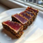 Four slices of perfectly seared tuna tataki, crusted with black and white sesame seeds, resting on a white plate.