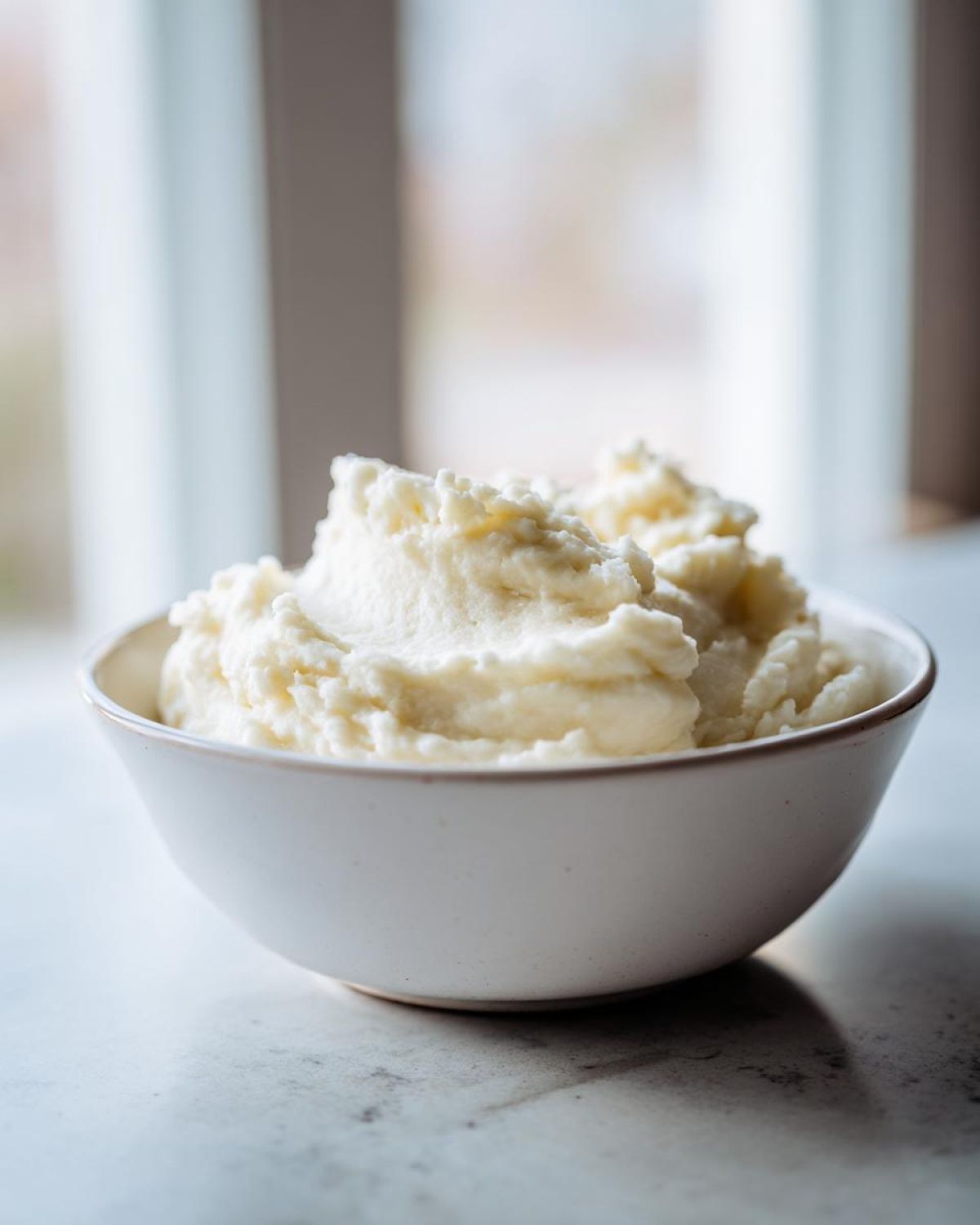 A white bowl filled with light, fluffy mashed potatoes, set against a bright, softly lit background.