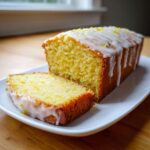 A loaf of moist Vegan Lemon Olive Oil Cake, partially sliced, drizzled with white lemon glaze and topped with zest.
