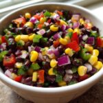 Close-up of a white bowl filled with colorful Cowboy Caviar featuring black beans, corn, red onion, and peppers.