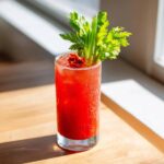 A tall, frosty glass of a vibrant red Kimchi Cocktails Recipe garnished with fresh celery and sun-dried tomatoes.