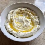 A white bowl filled with creamy whipped feta dip, swirled and drizzled generously with golden olive oil.