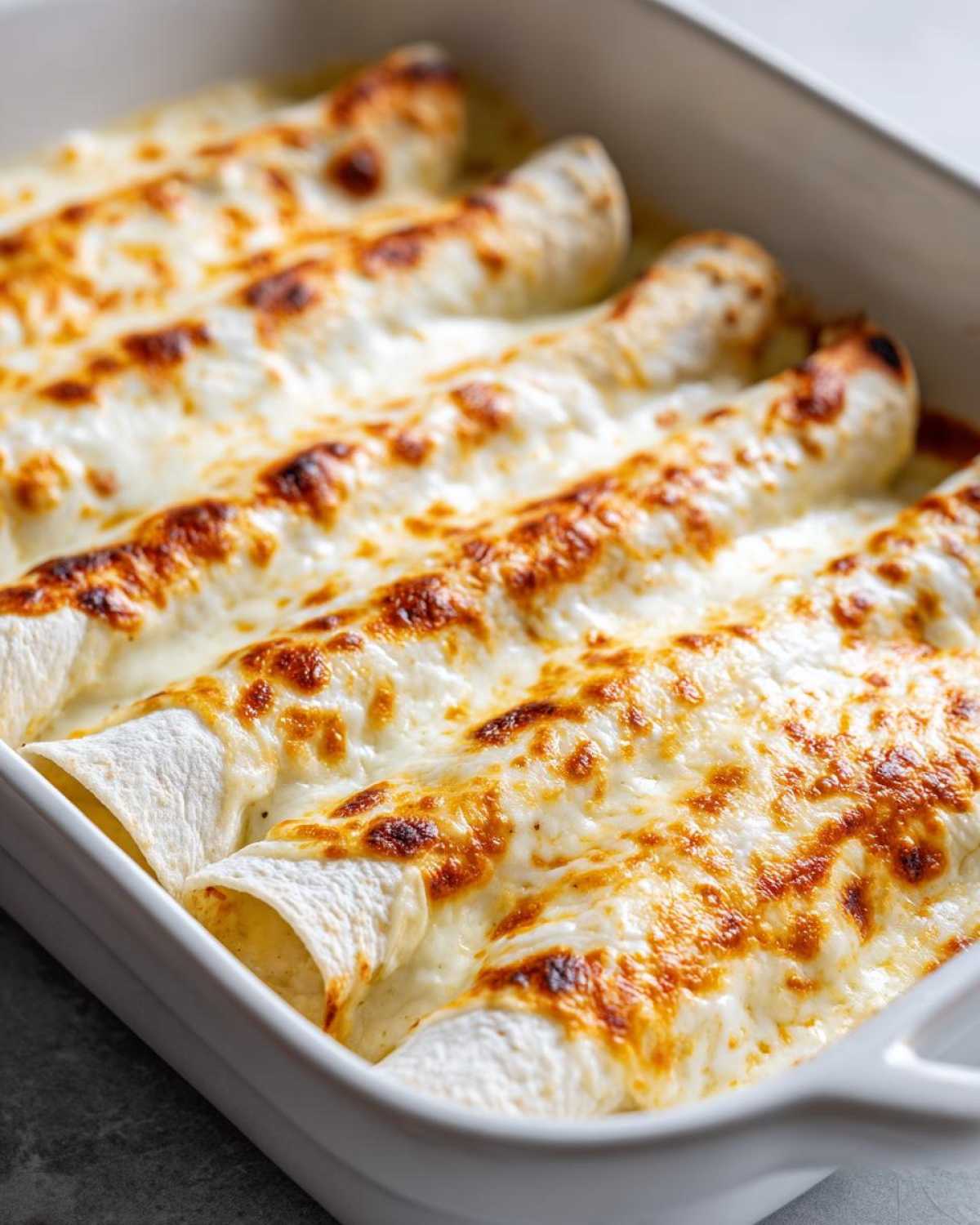Close-up of baked white chicken enchiladas covered in bubbly, browned cheese sauce in a white casserole dish.