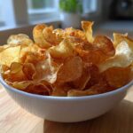 A white bowl overflowing with golden, crispy Air Fryer Potato Chips sitting on a wooden surface.