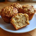 Close-up of four golden brown Banana Oatmeal Muffins on a white plate, one is cut in half showing the moist interior.