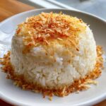A perfectly molded mound of white coconut rice topped and surrounded by crispy, toasted coconut flakes.
