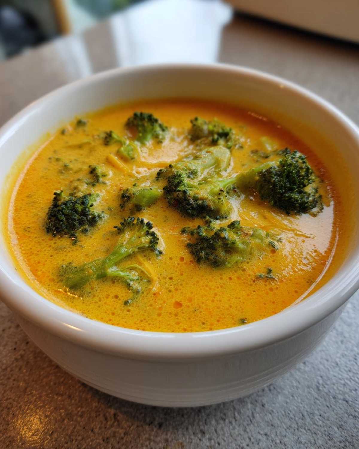 Close-up of creamy, orange-yellow Broccoli and Cheddar Soup with visible green broccoli florets in a white bowl.