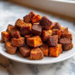 A large pile of perfectly cooked, seasoned, and Crispy Roasted Sweet Potatoes cubes stacked on a white plate.