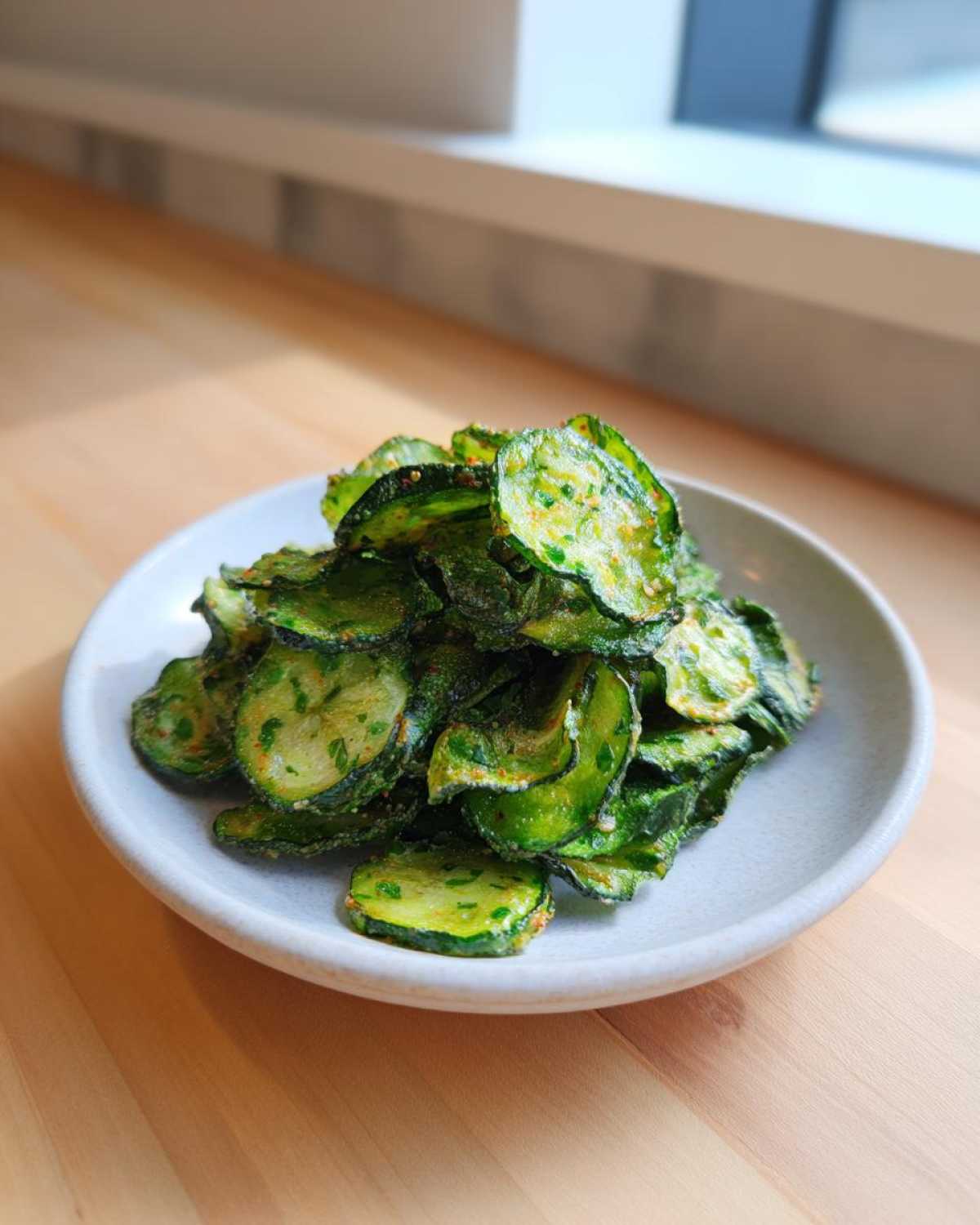 A small white bowl piled high with green, seasoned Crunchy Ranch Cucumber Chips on a light wooden surface.