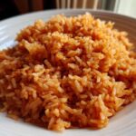 Close-up of a generous serving of fluffy, orange-red Mexican Rice piled high on a white, ridged plate.