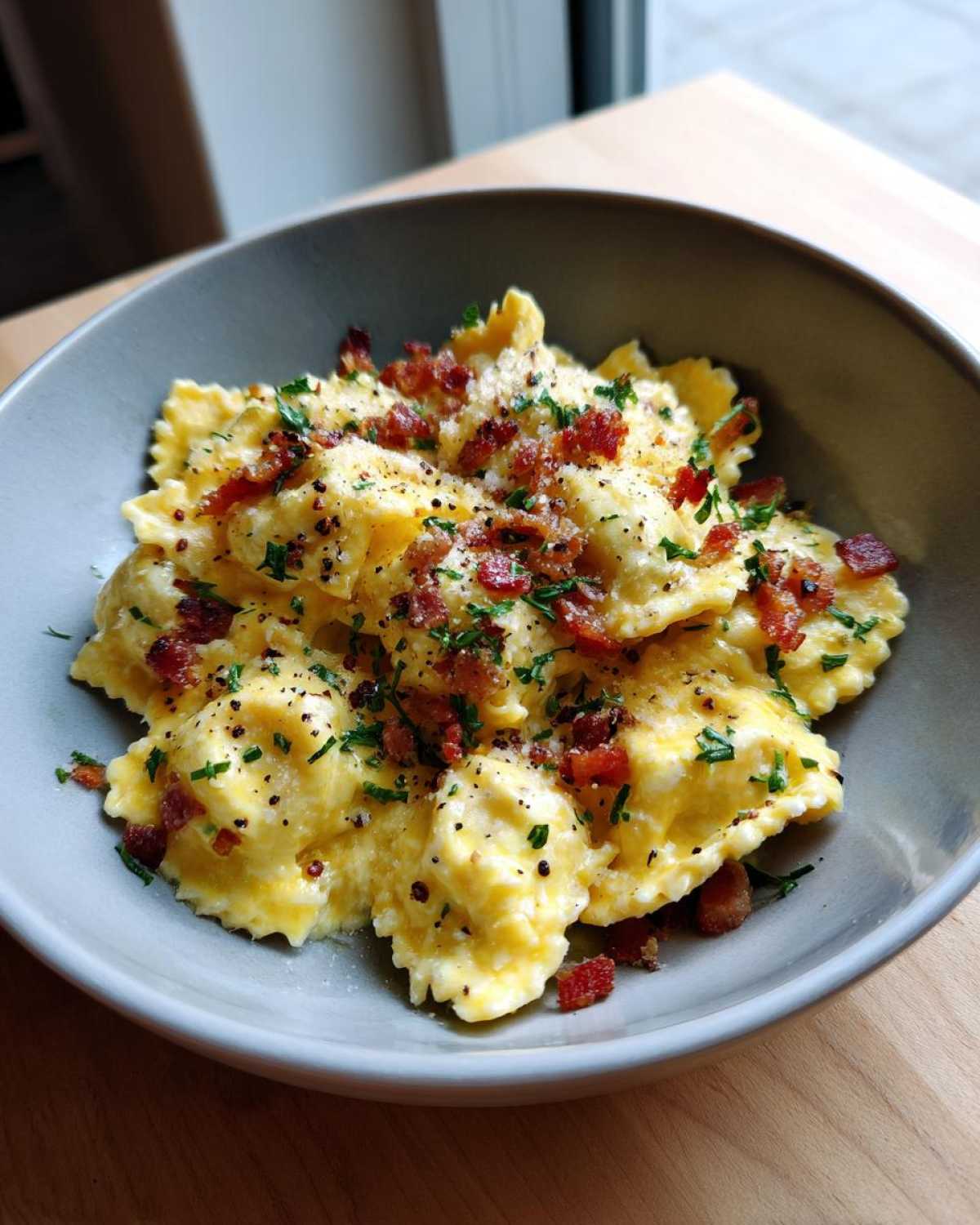 A close-up of creamy Garlic Ravioli Carbonara Pasta topped with bacon bits, parsley, and black pepper in a grey bowl.