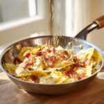 A stainless steel skillet filled with creamy Garlic Ravioli Carbonara Pasta topped with crispy bacon bits as cheese is sprinkled on top.