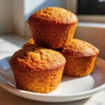 A stack of three golden brown Greek Yogurt Banana Muffins resting on a white plate, beautifully lit by sunlight.
