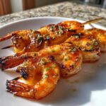 Close-up of glazed Sweet Chili Grilled Bang Bang Shrimp Skewers on a white plate, glistening with sauce and herbs.