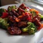 Close-up of Teriyaki Chicken Stir Fry featuring glazed chicken pieces, bright green broccoli, and red peppers on a white plate.